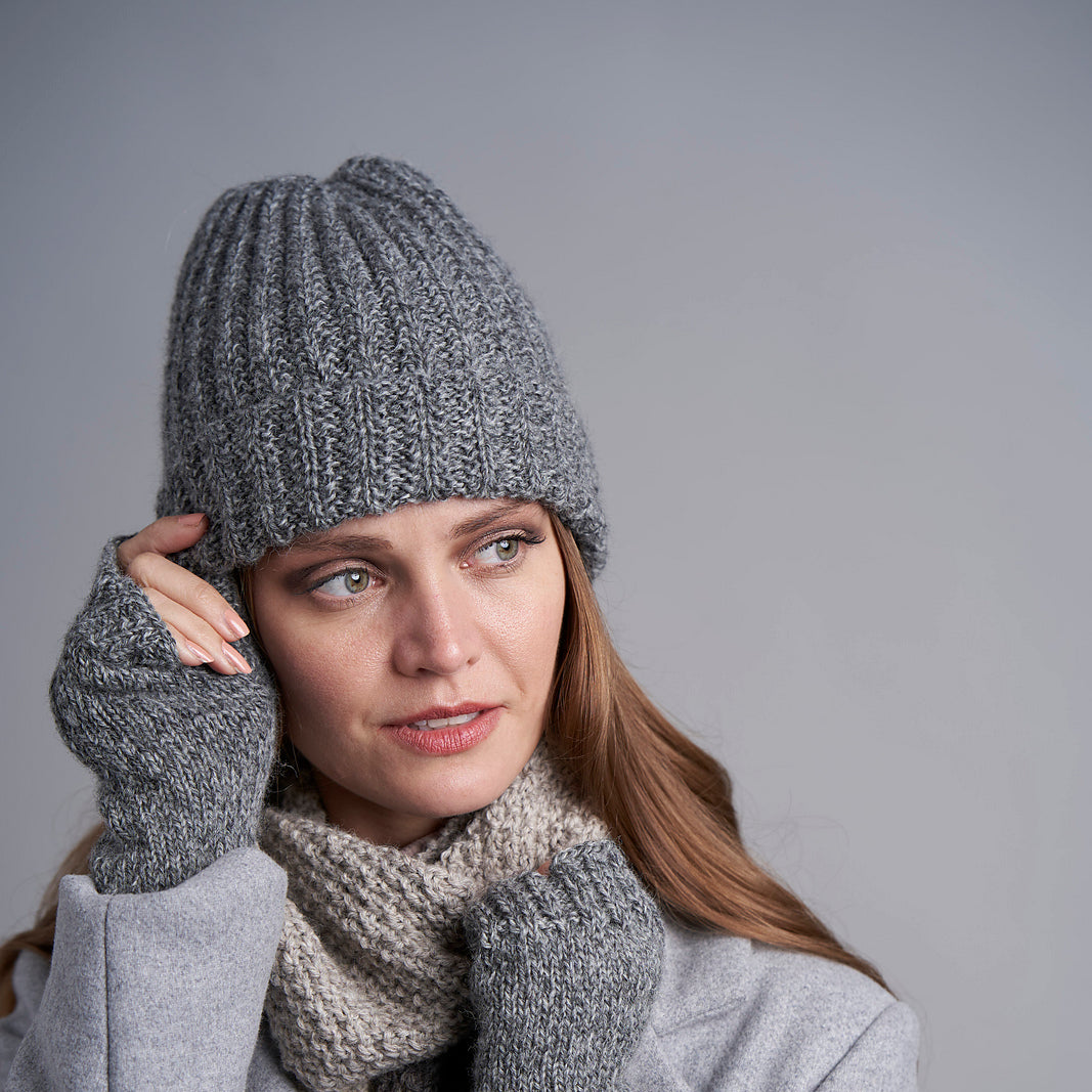 A woman wearing a gray ribbed knit beanie, matching gray knit fingerless gloves, a beige knit scarf, and a light gray coat, posed against a solid gray studio background.