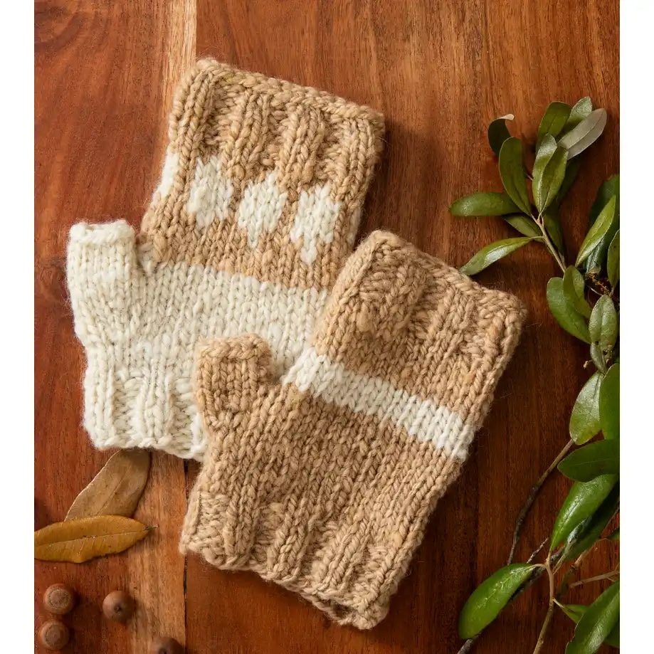 A pair of chunky knit fingerless gloves in natural beige and cream, styled on a warm wooden surface with green leaves and small acorns.