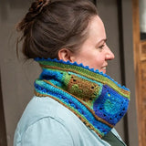 A woman in profile wearing a chunky hand-crocheted cowl neck warmer with a multicolor granny square pattern in shades of blue, green, and brown.